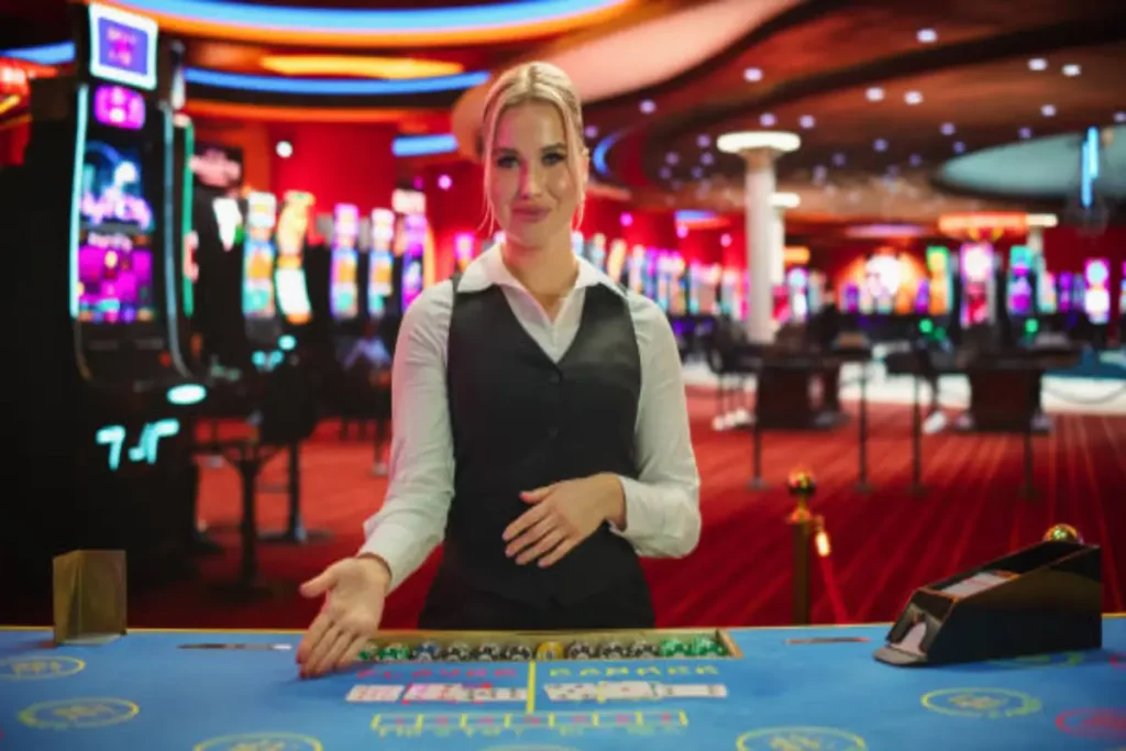 A casino dealer stands behind a gaming table, smiling, with slot machines in the background