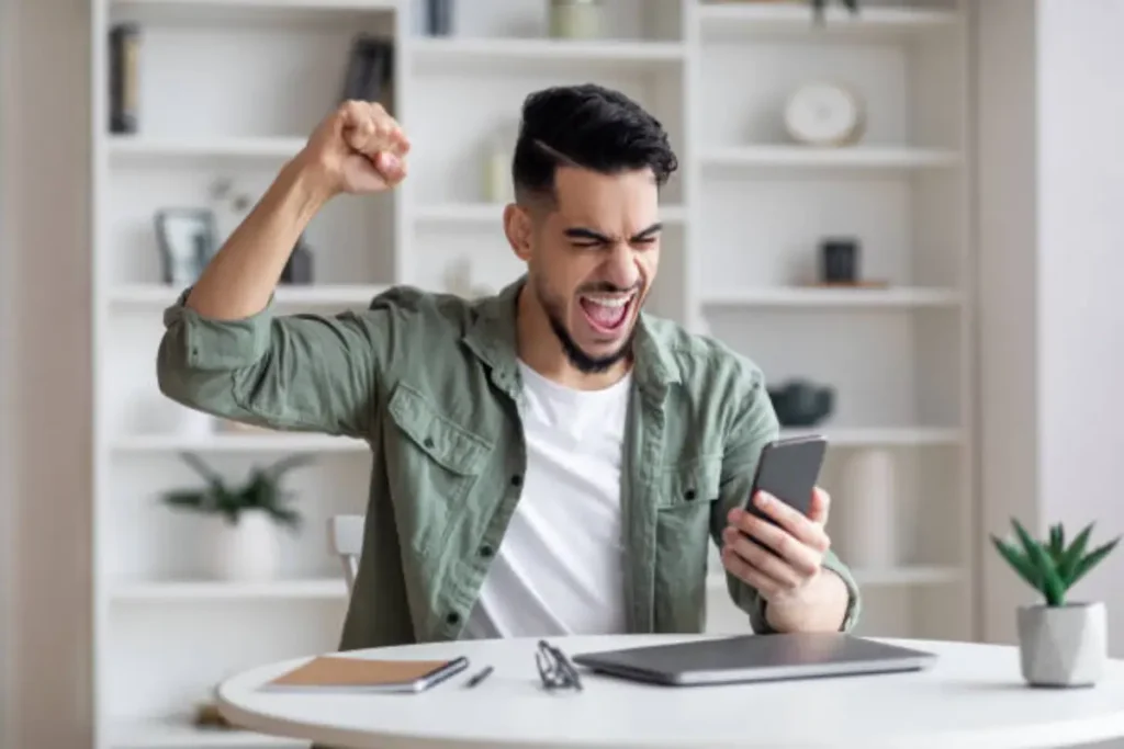 A man in a green shirt celebrates joyfully while holding a smartphone