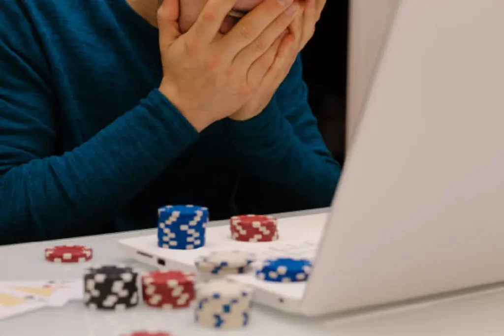 A person in a blue sweater sits at a laptop, hands over face, surrounded by poker chips