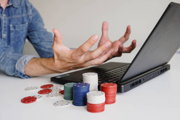 A person in a denim shirt gestures frustratedly in front of a laptop with stacks of colorful poker chips on the table