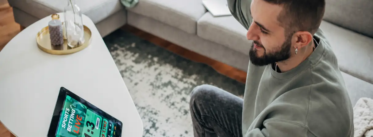A man sits on a couch looking excitedly at a tablet on a table displaying a sports betting app